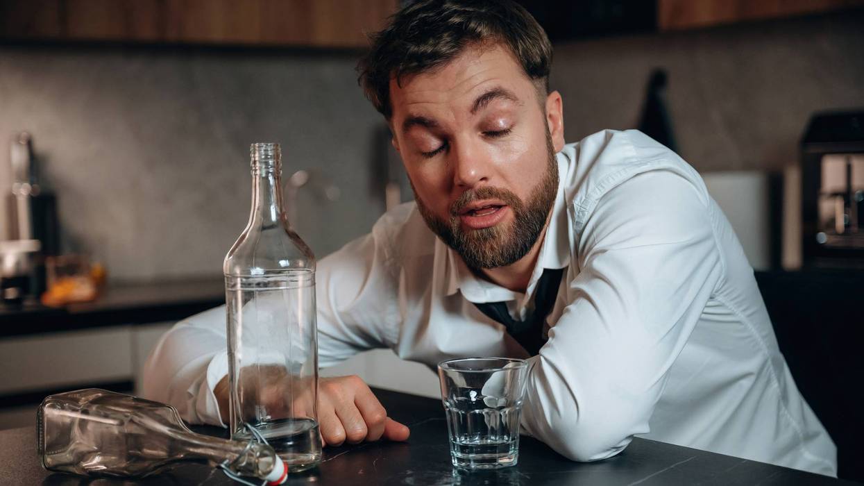 Man in disheveled attire appears intoxicated at a kitchen table, surrounded by empty bottles and a glass, illustrating the effects of excessive alcohol consumption