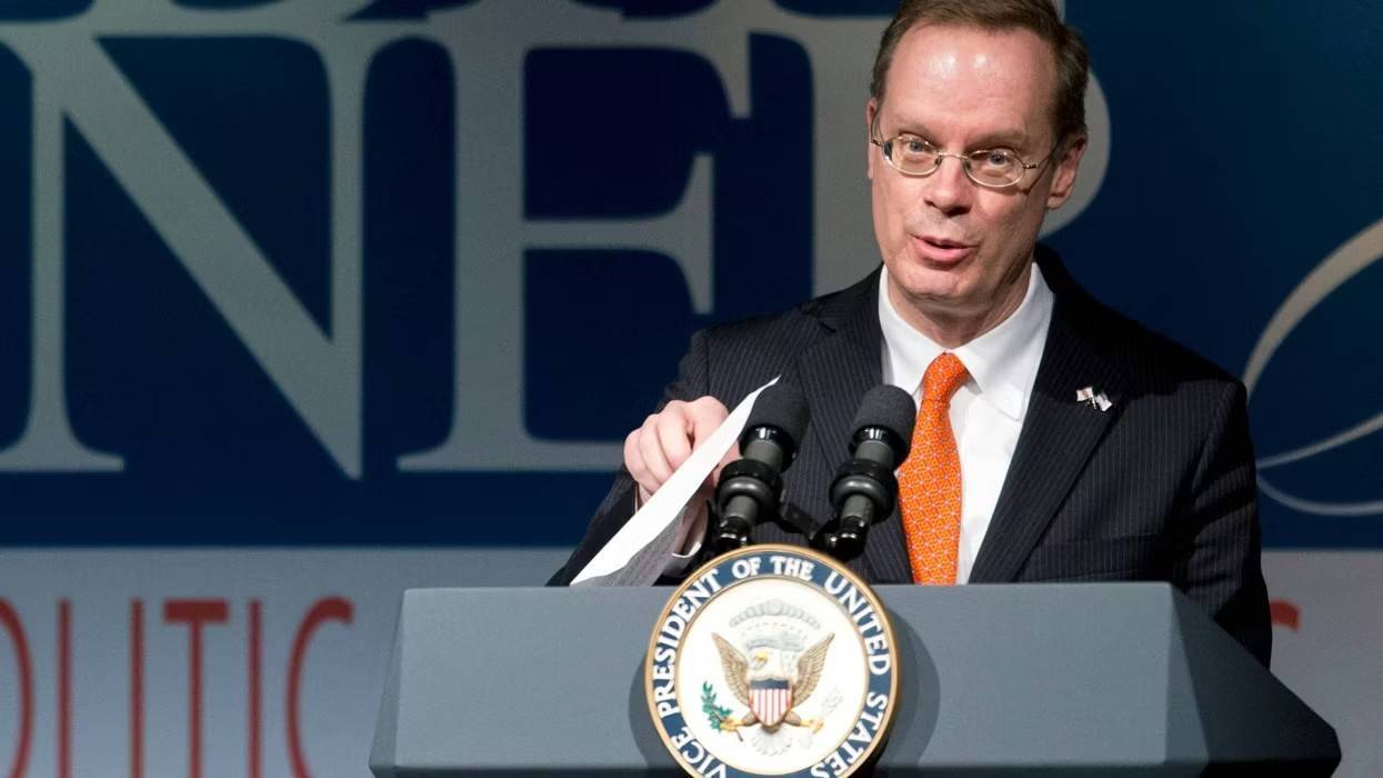 Man in suit, glasses, orange tie, speaks at podium with US Vice Presidential seal, holding papers.
