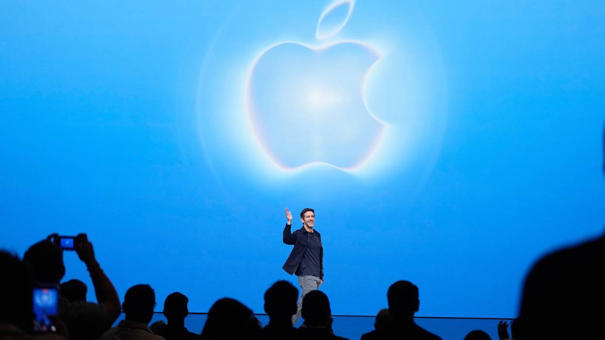 Man waves on stage in front of a large glowing Apple logo on a blue screen, audience below.