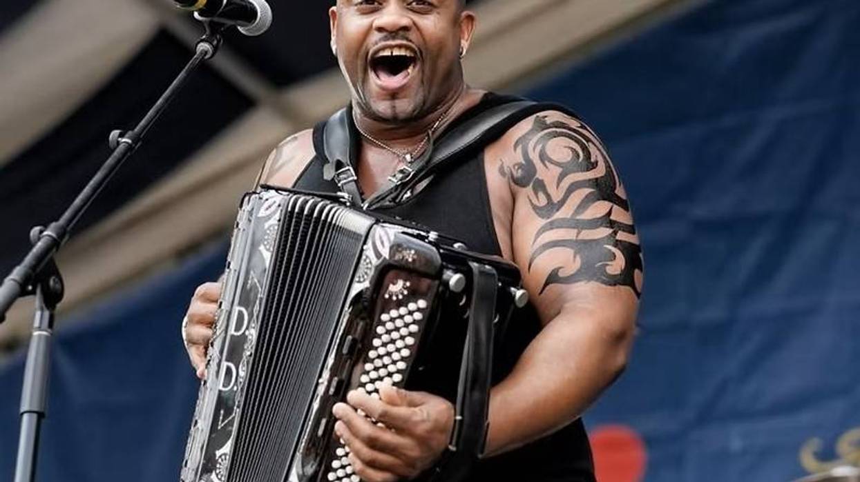 Man with mohawk and tattoos playing accordion, singing enthusiastically into mic.