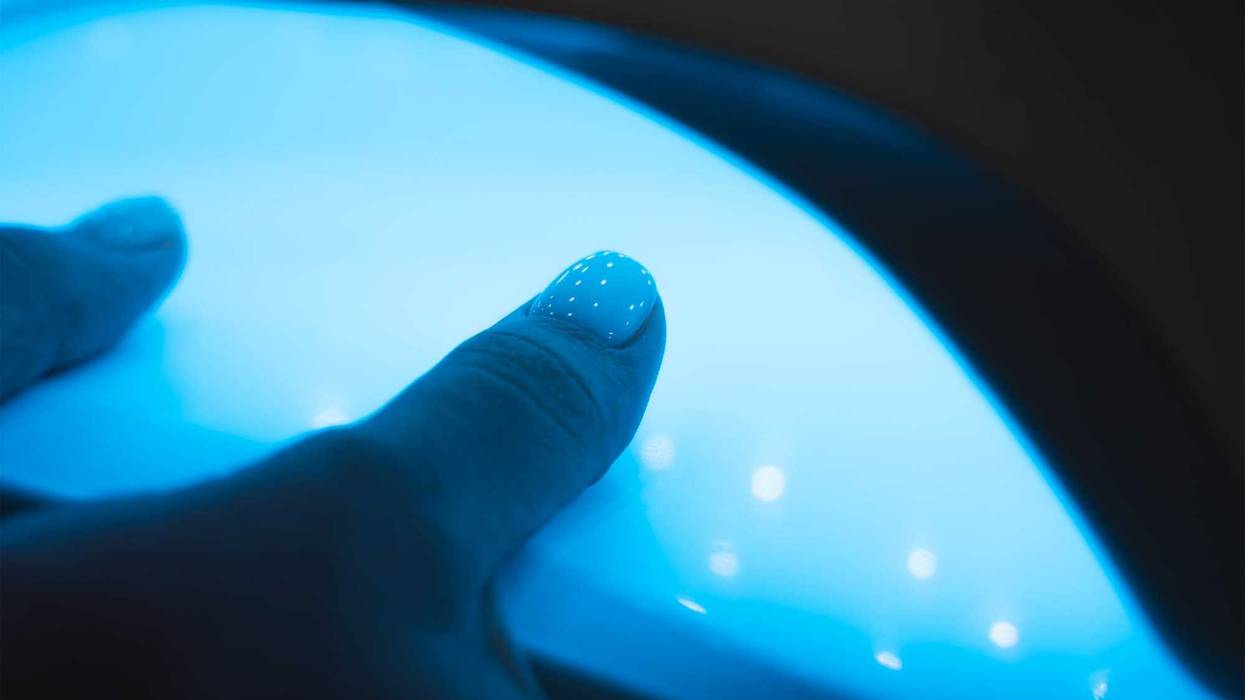 Manicurist master is covering painting client's nails top coat shellac, hands closeup. One hand drying in UV lamp. Professional manicure with gel polish. Nail art, hands care, beauty industry concept.