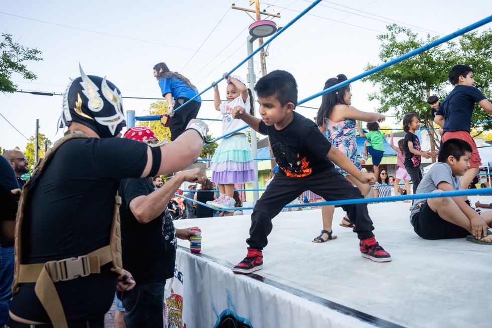 Masked Lucha Libre wrestler watches kids in an outdoor ring, one boy mimics a punch.