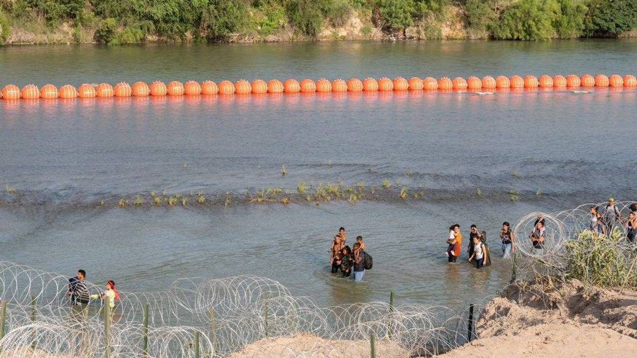 Migrants walk by a string of buoys placed on the water along the Rio Grande border with Mexico in Eagle Pass, Texas