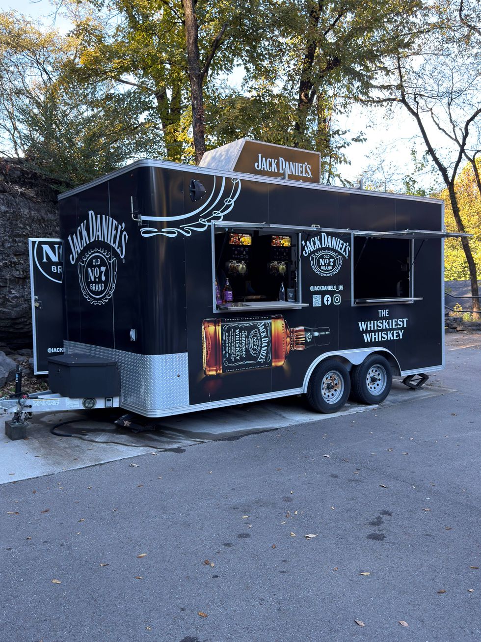 Mobile whiskey bar set in a serene outdoor scene with trees. The black trailer features Jack Daniel