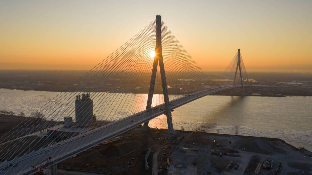 Modern cable-stayed bridge at sunrise, golden sun behind its main tower over an icy river.