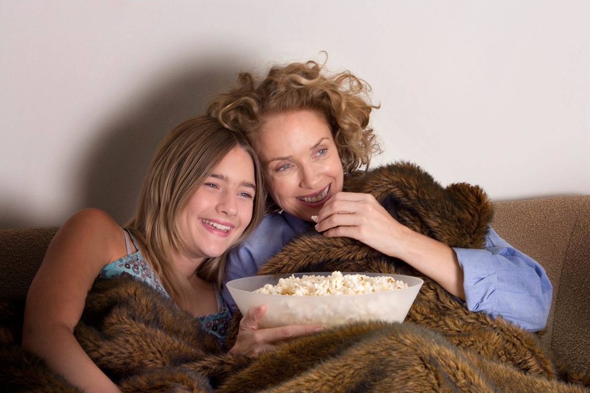 Mother and daughter (13-15) on sofa sharing popcorn, laughing