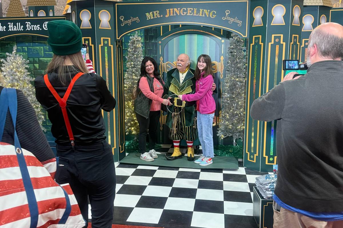 Mr. Jingeling at the Christmas Connection posing with two girls for a photo. Multiple people are taking pictures of them and they are in front of a green and gold decorative background.