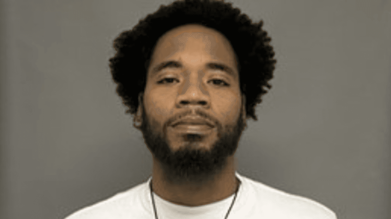 Mugshot of a Black man with an afro and beard, wearing a white t-shirt.