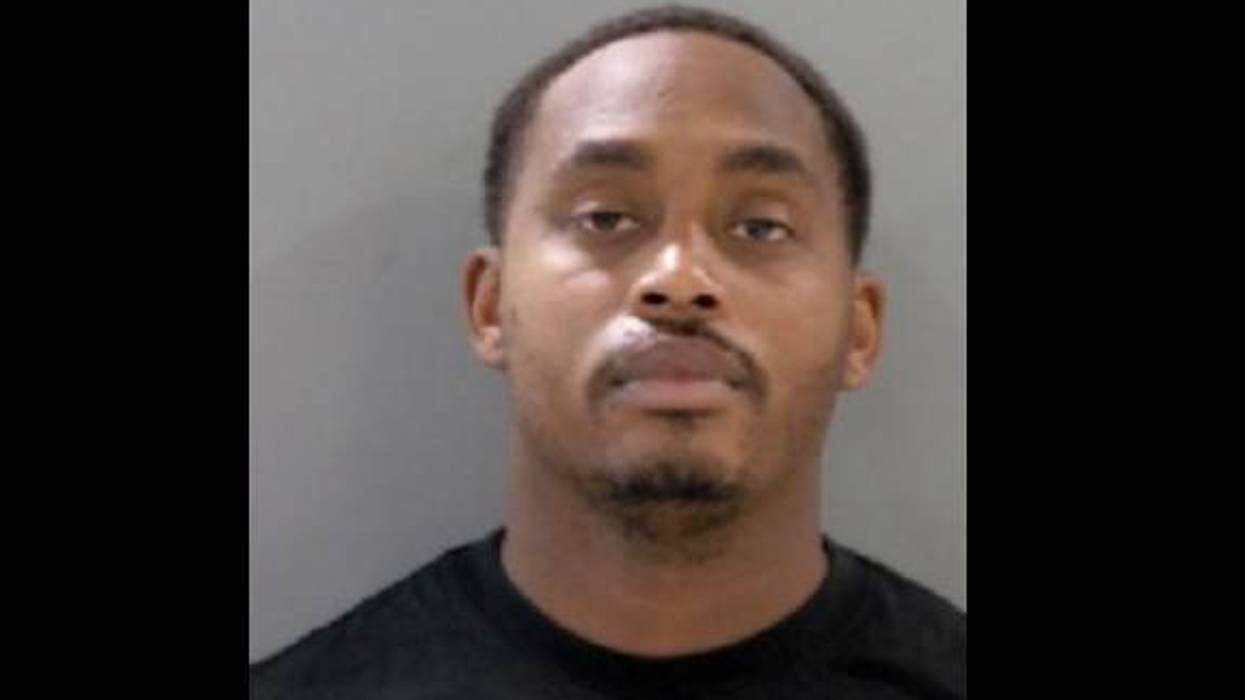 Mugshot of a Black man with short hair, a mustache, and a short beard, wearing a dark shirt.