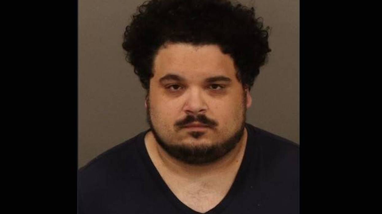 Mugshot of a man with dark, curly hair and a full beard, wearing a dark V-neck shirt.