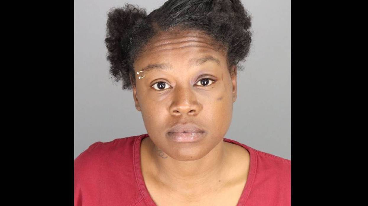Mugshot of a woman with dark skin, dark curly hair, an eyebrow piercing, and a red shirt.