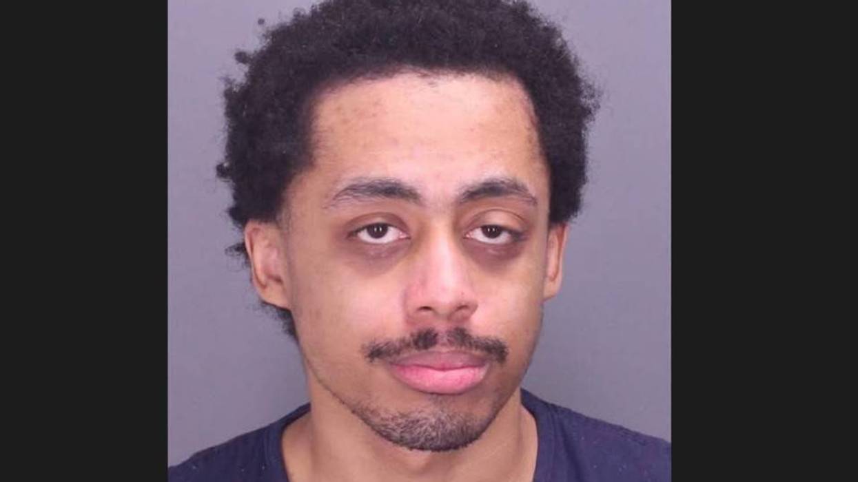 Mugshot of a young man with dark, curly hair, mustache, short beard, and dark circles under eyes.