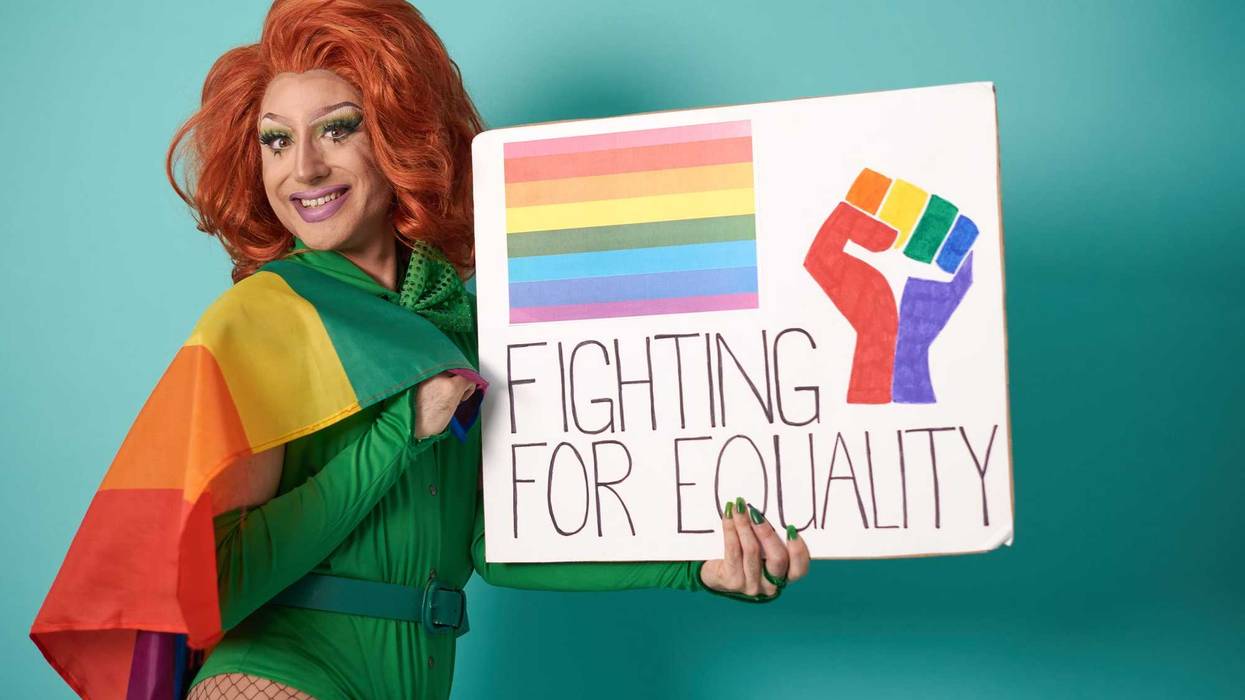 Optimistic transgender person with LGBT flag looking at camera with smile and showing placard with Fighting For Equality inscription against blue background