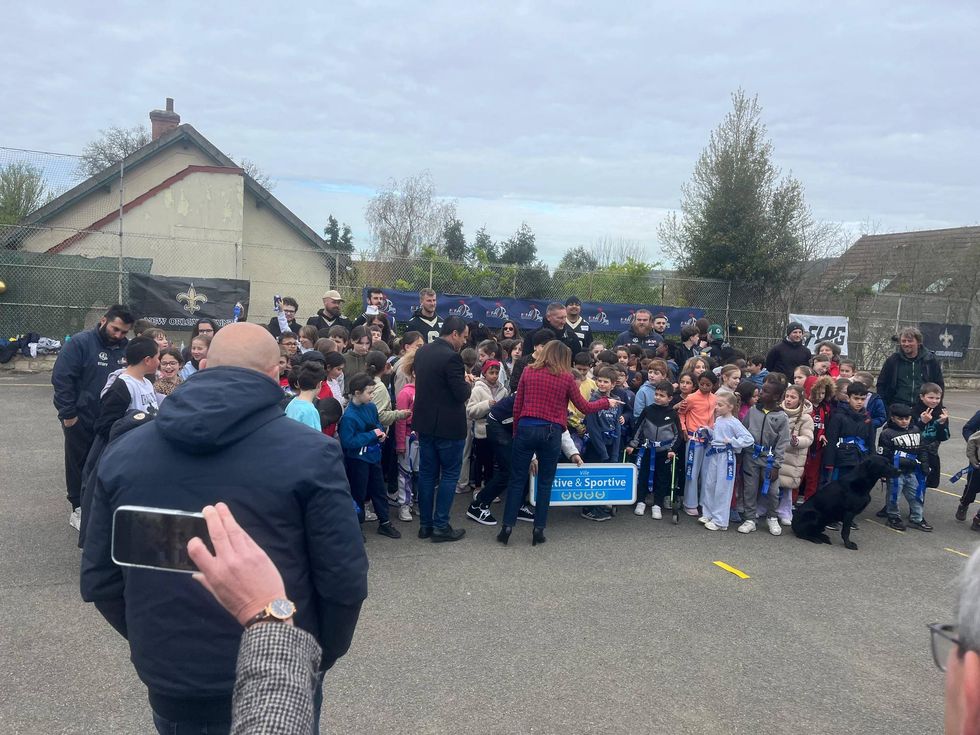 Outdoor gathering of children wearing flag football belts and adults with New Orleans Saints banners.
