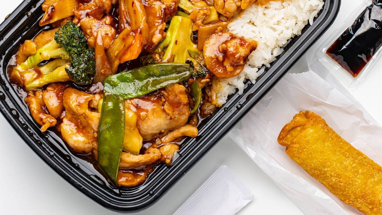 Overhead view of Chinese cuisine takeout meal with chicken stir fry, rice, egg roll, and soy sauce packets isolated on white backdrop.