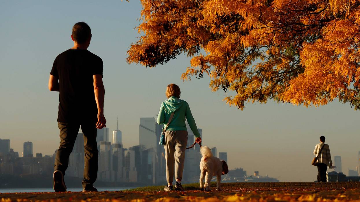 People walk along a walkway covered in fall foliage in Edgewater, New Jersey, as the sun rises over the New York skyline on Oct. 28, 2023, when highs hit 80 degrees in Central Park