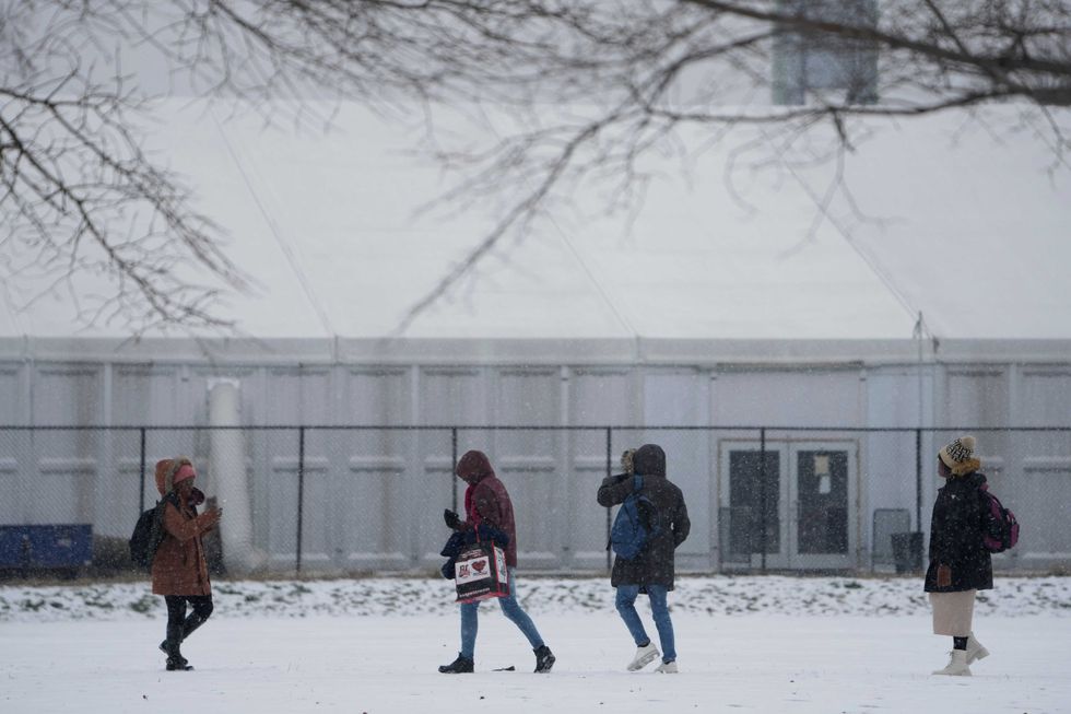 People walks across a snowy athletic field in front of a temporary shelter for migrants on Randall