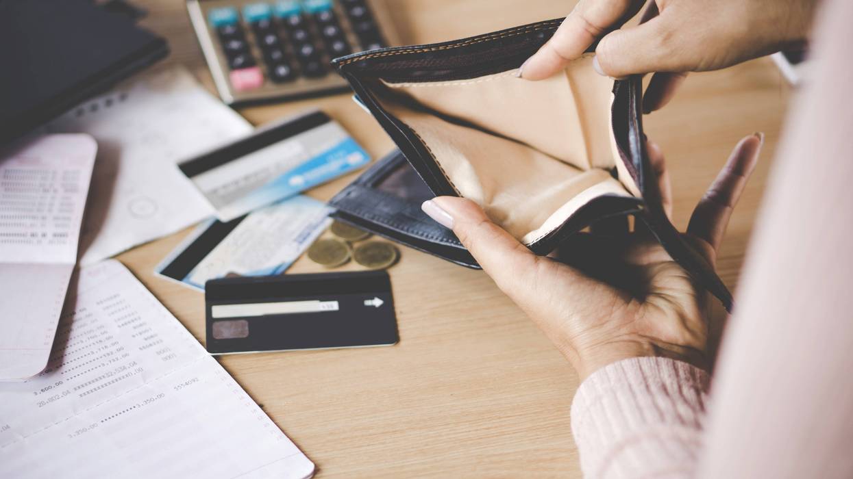 person opening wallet that's empty inside, background has a desk with bills, credit cards, coins, a calculator