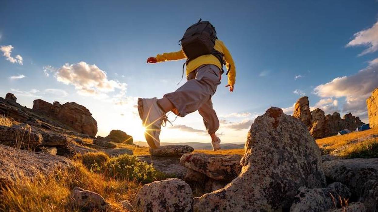 Person with backpack leaping over rocks at sunset. Sun flare on boot, distant tents.