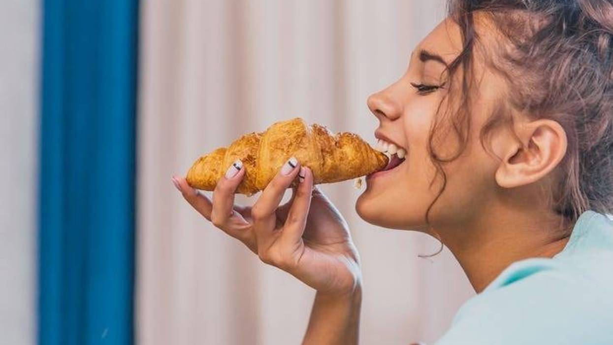 Photo of a cheerful young woman happy. Going to the table and eating croissant.