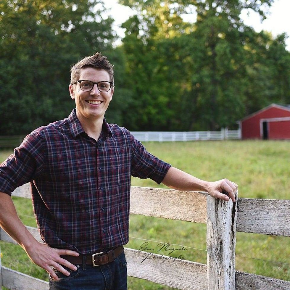 Photo of man in 30s holding onto wooden fence, smiling with glasses, and wearing flannel