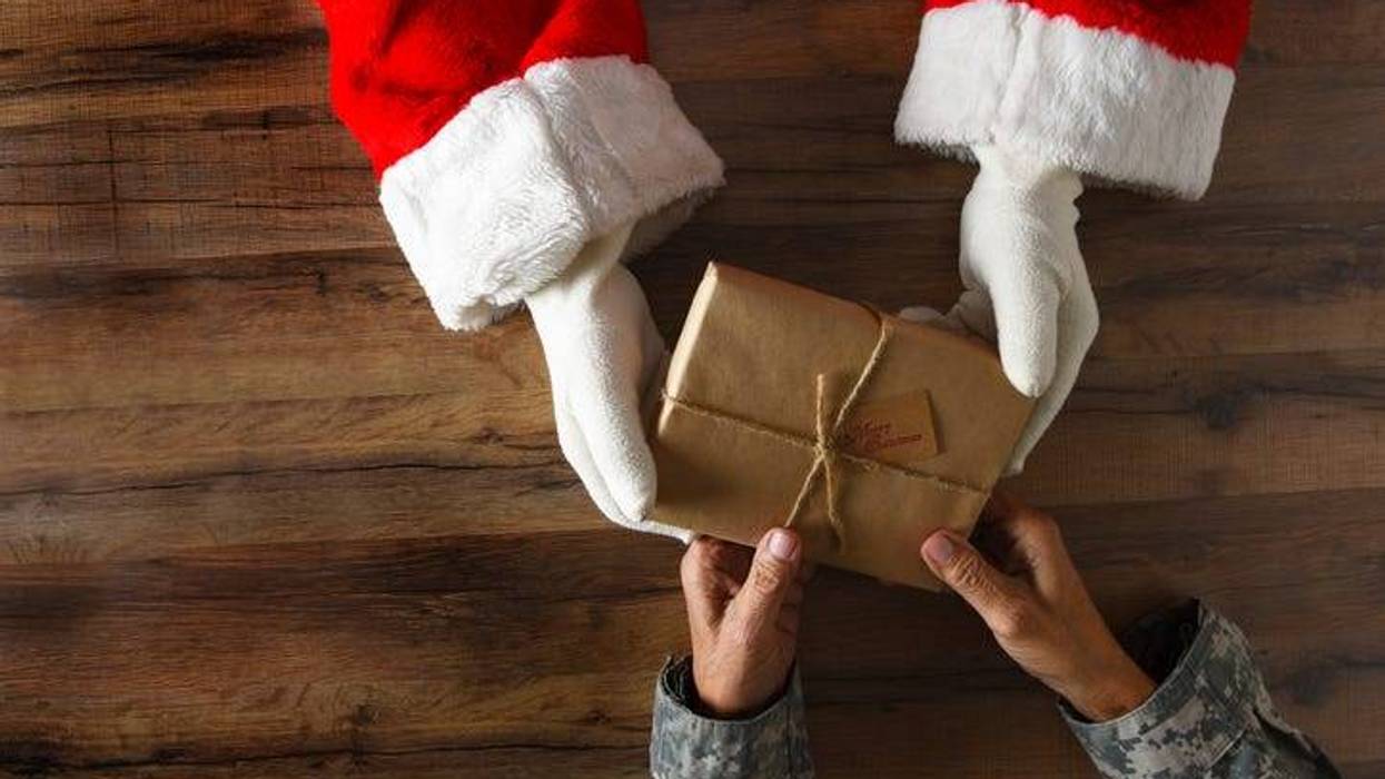 Photo of Santa handing a plainly wrapped present to a soldier. Only their hands holding the gift are visible.