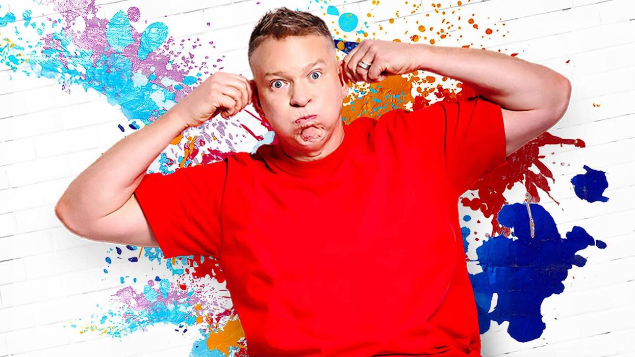 Playful man in red shirt making a funny face and pulling ears on a colorful paint-splattered wall.