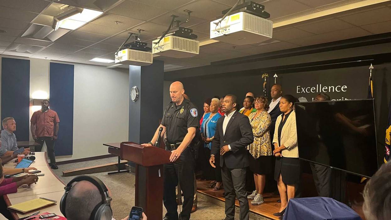 Police chief, with the Mayor, the Superintendent, and a small crowd of local leaders, addresses a room full of reporters