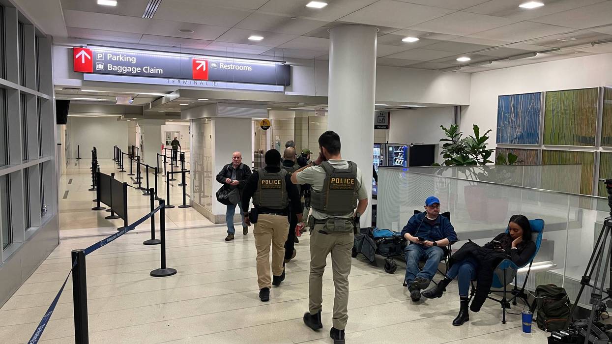 Police in vests walk through an airport terminal past people sitting in chairs near a camera setup.