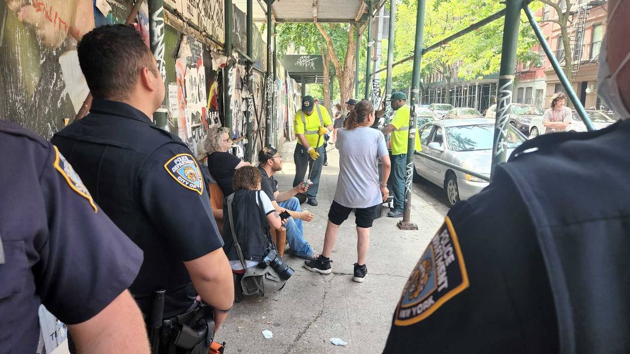 Police watch as encampment resident Johnny Grima, another resident, a neighbor and a news photographer huddle around an arm chair — the last piece of the encampment after the tents, bedding and other belongings were thrown away. The NYPD would go on to physically remove them from the chair, which sanitation workers then destroyed.