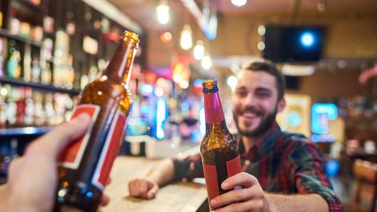 Portrait of happy bearded man clinking beer bottles with friends while having fun in pub sitting at bar counter, focus on foreground