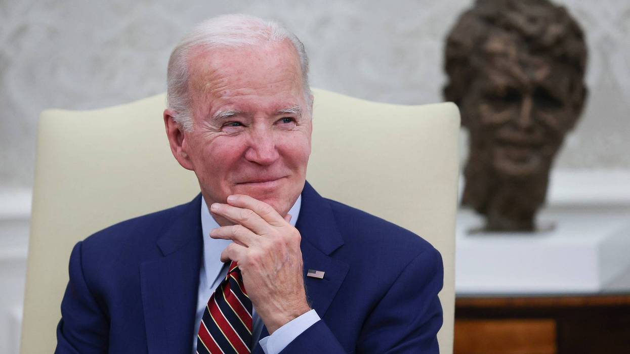 president joe biden sitting on a cream colored chair wearing blue suit holding his hand up to his chin