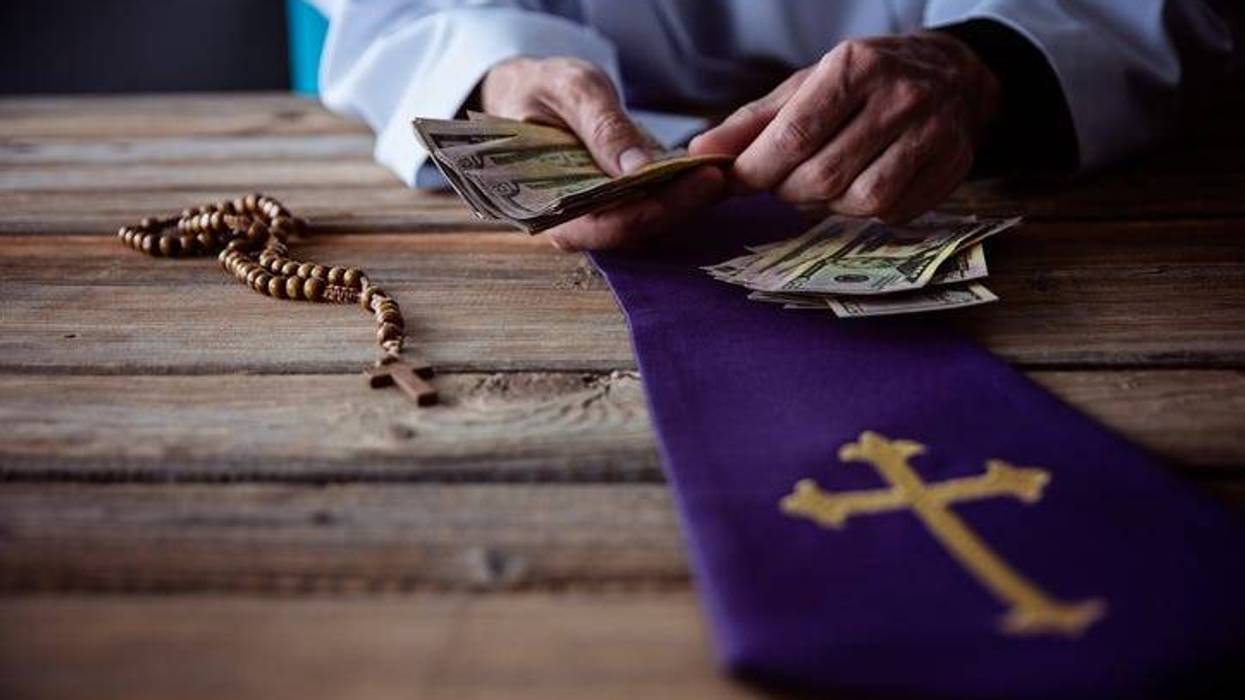 Priest shown counting money on a wooden table. A rosary and mantle are also displayed on the table.