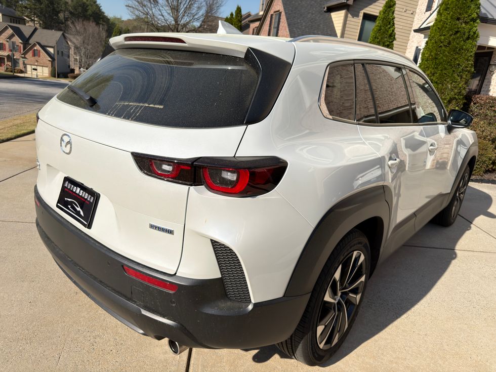 Rear-side view of a white Mazda CX-50 Hybrid SUV on a sunny suburban driveway.