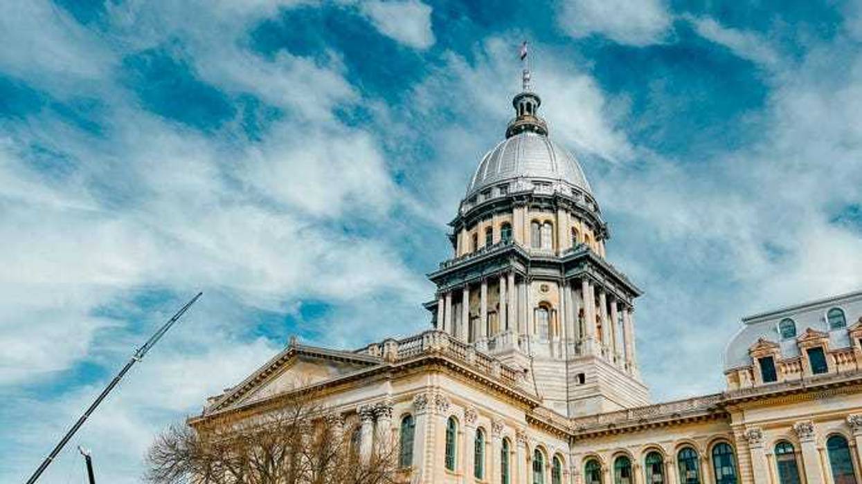 Rear views of the Illinois State Capitol Building in Springfield, Illinois, USA. Cloudy blue skies overhead. The US flag, State of Illinois Flag, and Black History Month Flag fly atop the dome. Construction crane in the view as construction work continues.