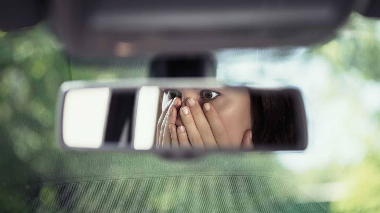 Reflection of young woman with frightened eyes covering her face with hands in the car rear view mirror.
