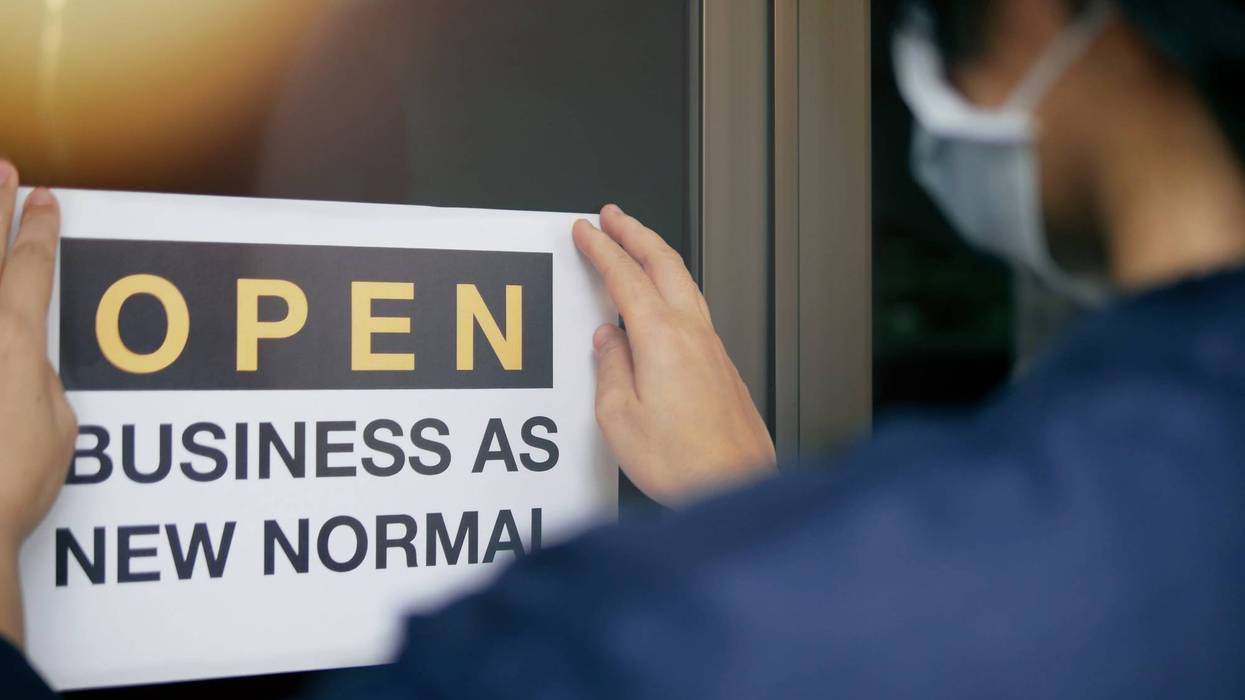 Reopening for business adapt to new normal in the novel Coronavirus COVID-19 pandemic. Rear view of business owner wearing medical mask placing open sign "OPEN BUSINESS AS NEW NORMAL" on front door.