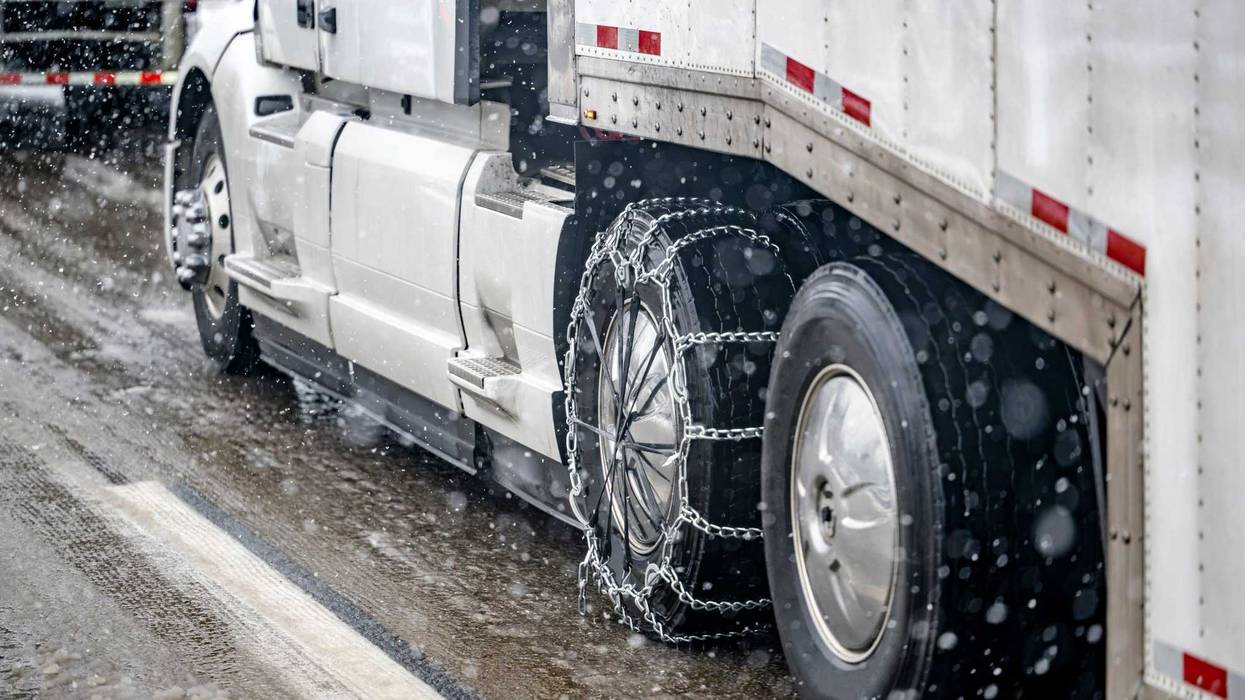 Required chains for better grip on slippery road surfaces on a white big rig semi truck drive axle wheel transporting cargo in dry van semi trailer running at winter weather during a snow storm