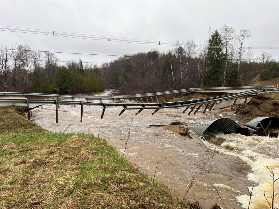 Road and culverts washed out by a raging brown river, leaving broken guardrails.