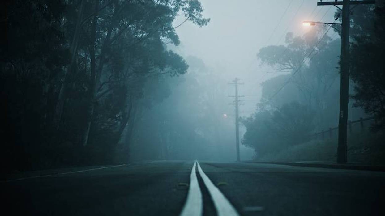 Road with double white lines disappearing into thick fog, flanked by dark trees & a street light.