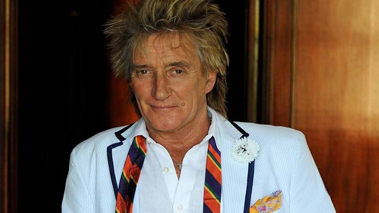 Rod Stewart, spiky blonde hair, smiling in a light blue striped blazer, colorful tie, and white boutonniere.