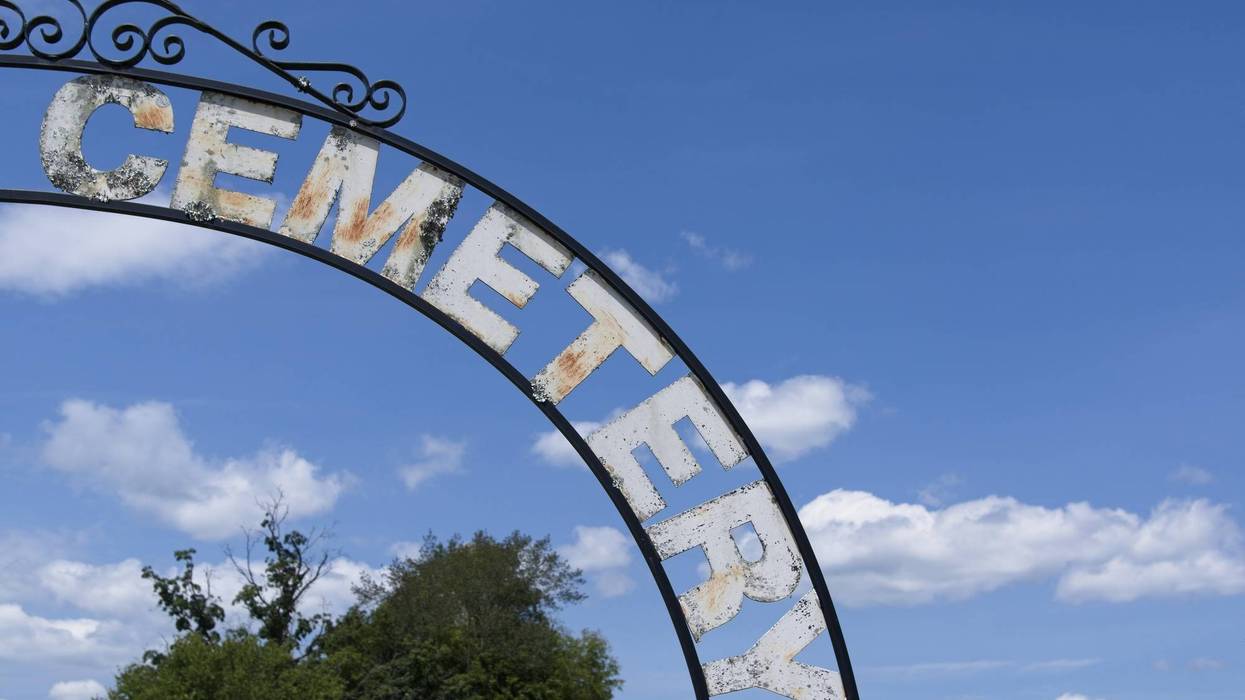 Rustic "CEMETERY" sign arch against blue sky with clouds and trees.