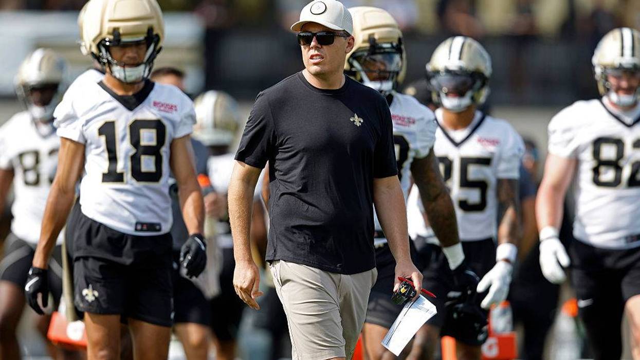 Saints coach in cap and sunglasses walks with players in white jerseys on the field.