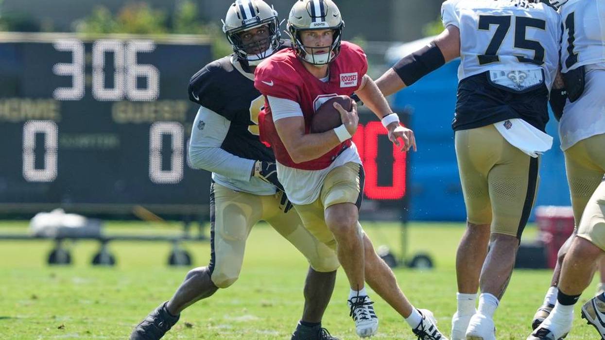 Saints QB in red jersey scrambling with football, pursued by a defender at practice.