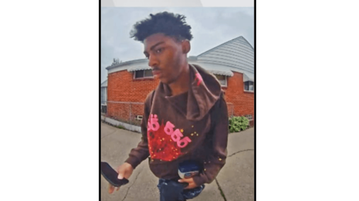 Security image of young Black man in brown '555' hoodie, holding phone & object, outside house.