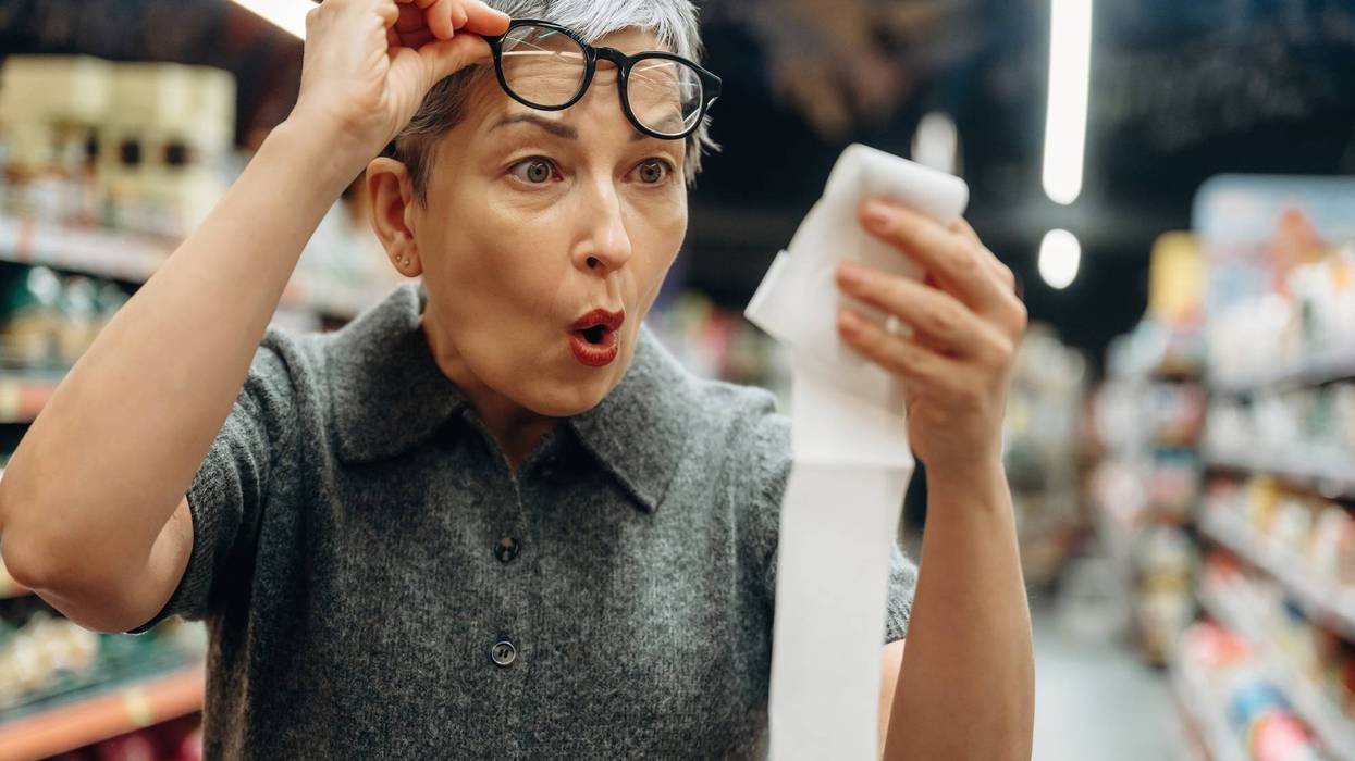 Senior woman with short gray hair wearing glasses looks surprised while holding a long receipt inside a grocery store aisle filled with products