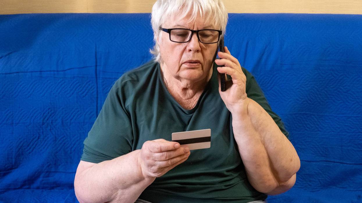 Serious old woman in glasses sitting on sofa holding credit card and talking on phone. Concept of scam, money loss, banking protection, finance fraud, customer service, remote payment