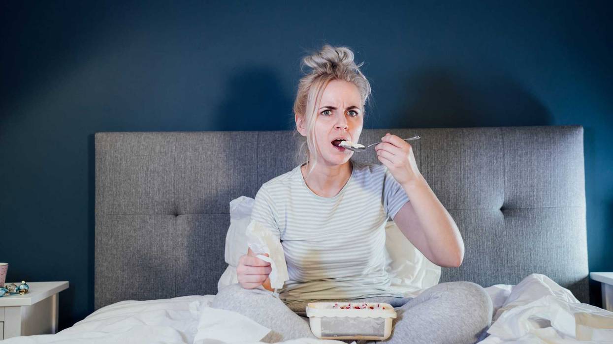 Shocked, stressed, surprised woman eating ice cream, holding a remote control while watching TV in bed. Scary horror movie or terrible news, scare in her eyes Overeating, Jamming of negative emotions.