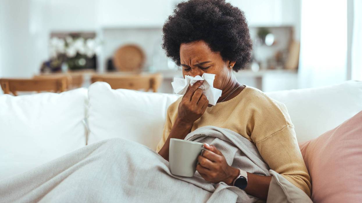 Sick young woman sitting on sofa blowing her nose at home in the sitting room. Photo of sneezing woman in paper tissue. Picture showing woman sneezing on tissue on couch in the living-room