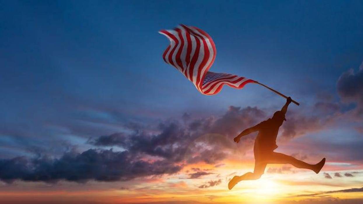 Silhouette of man jumping while holding US American flag in front of beautiful sunrise.